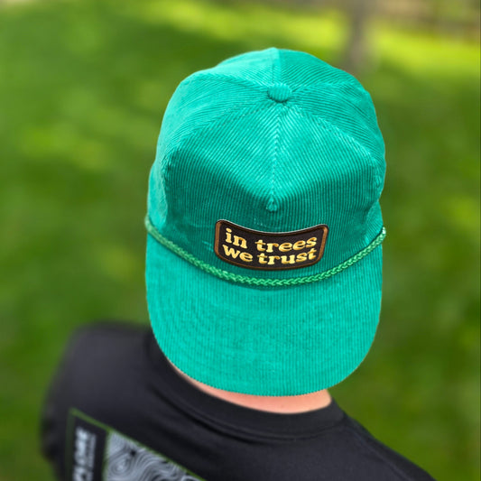 A green corduroy baseball cap with a brown and gold 'in trees we trust' patch, worn by a person in a black shirt against a blurred green outdoor background.