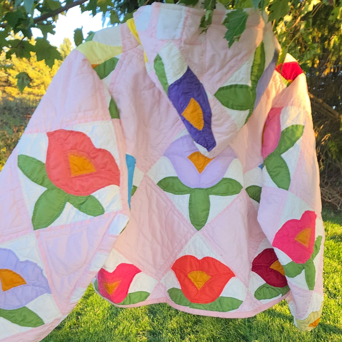 A light pink quilted jacket with large red, purple, and yellow floral appliques hanging from a tree branch outdoors on a green lawn.