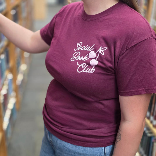 A woman in a library aisle wearing a maroon t-shirt with a pink graphic that reads 'Social Media Club', paired with blue jeans.